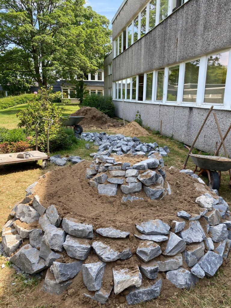 Bau eines Steinhügels mit Erde und Steinen neben einem Schulgebäude.