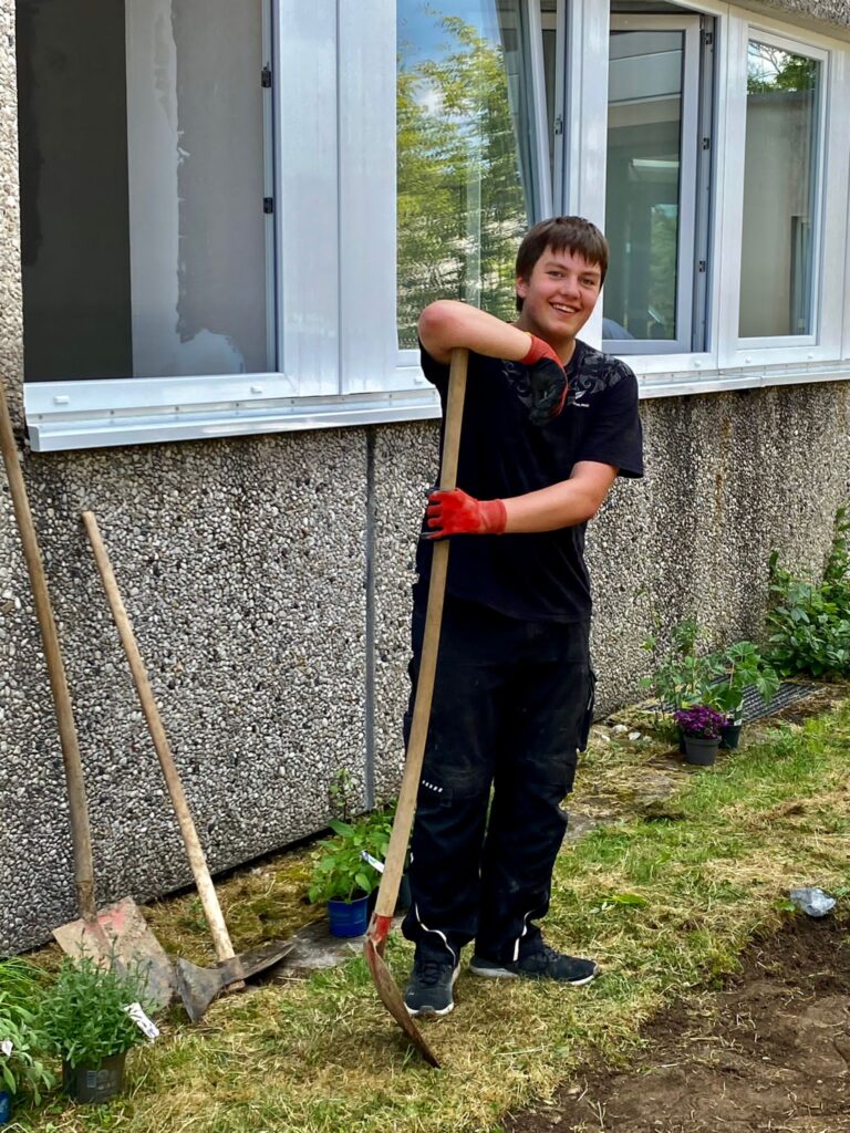 Schüler steht mit einer Schaufel in der Hand neben einem Fenster und lächelt.