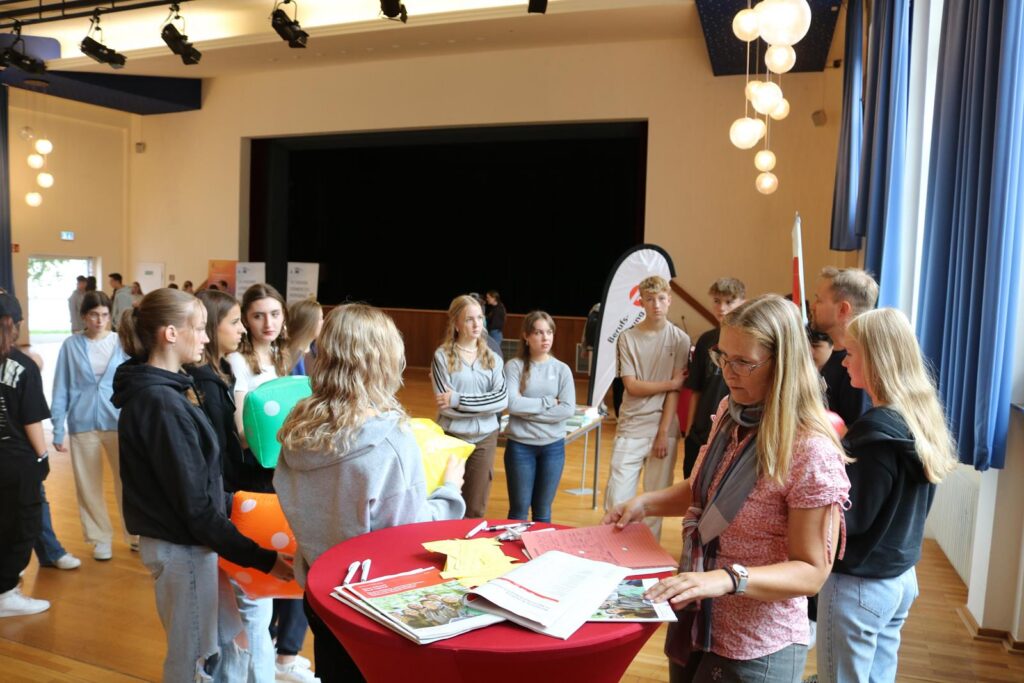 Schüler stehen in einem Raum um Tische, Gespräche und Aktivitäten finden statt.