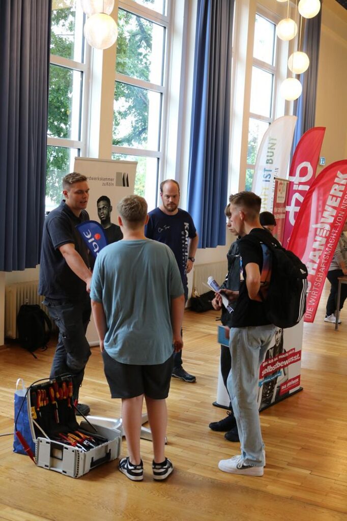 Leute diskutieren an einem Informationsstand in einem Raum mit großen Fenstern.