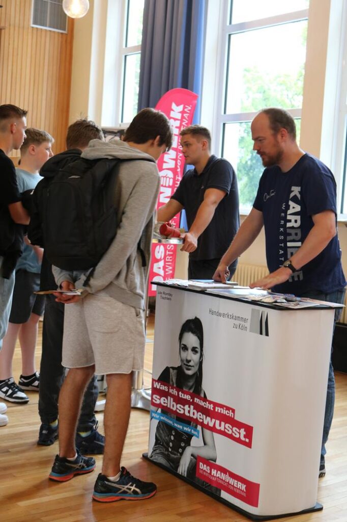 Zwei Männer stehen an einem Stand, helfen Schülern bei Informationen.
