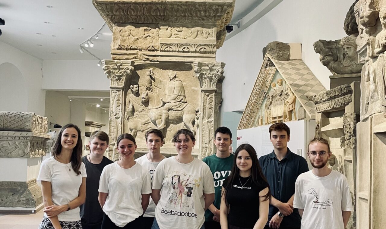 Gruppe von Schülern steht in einem Museum vor einer steinernen Säule.