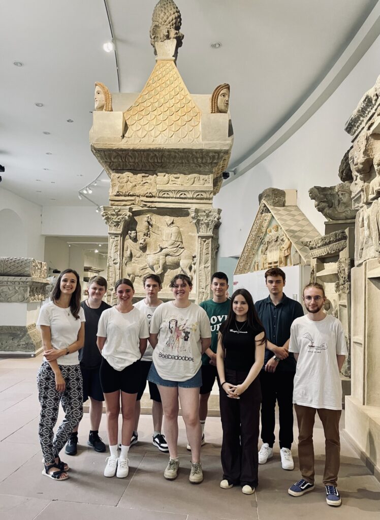 Gruppe von Schülern steht in einem Museum vor einer steinernen Säule.