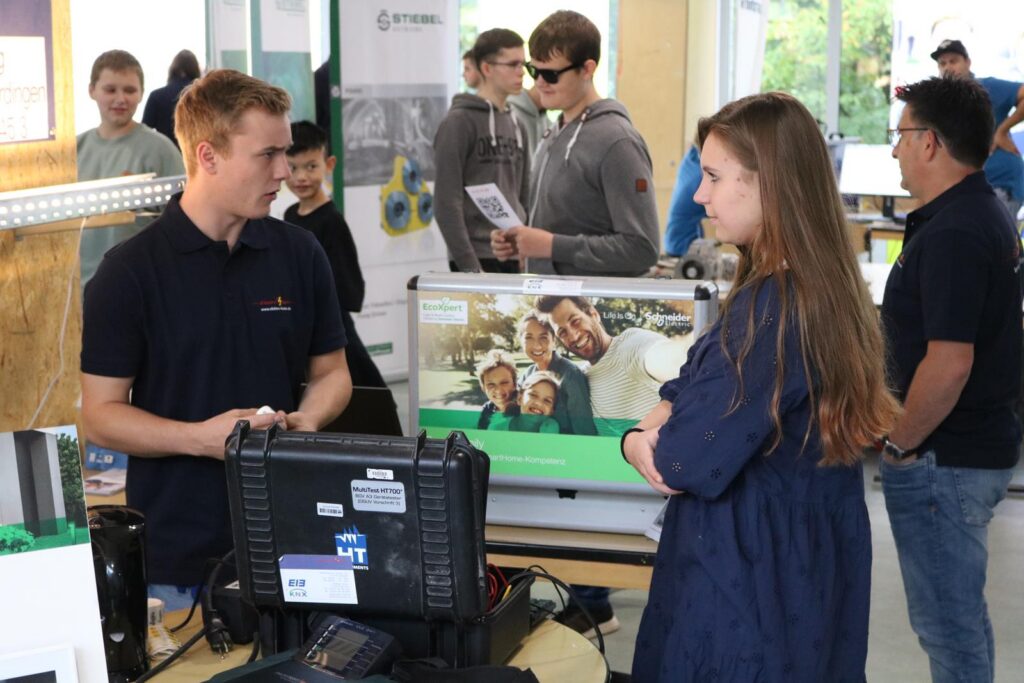 Zwei Personen stehen an einem Stand, Gespräche finden im Hintergrund statt.