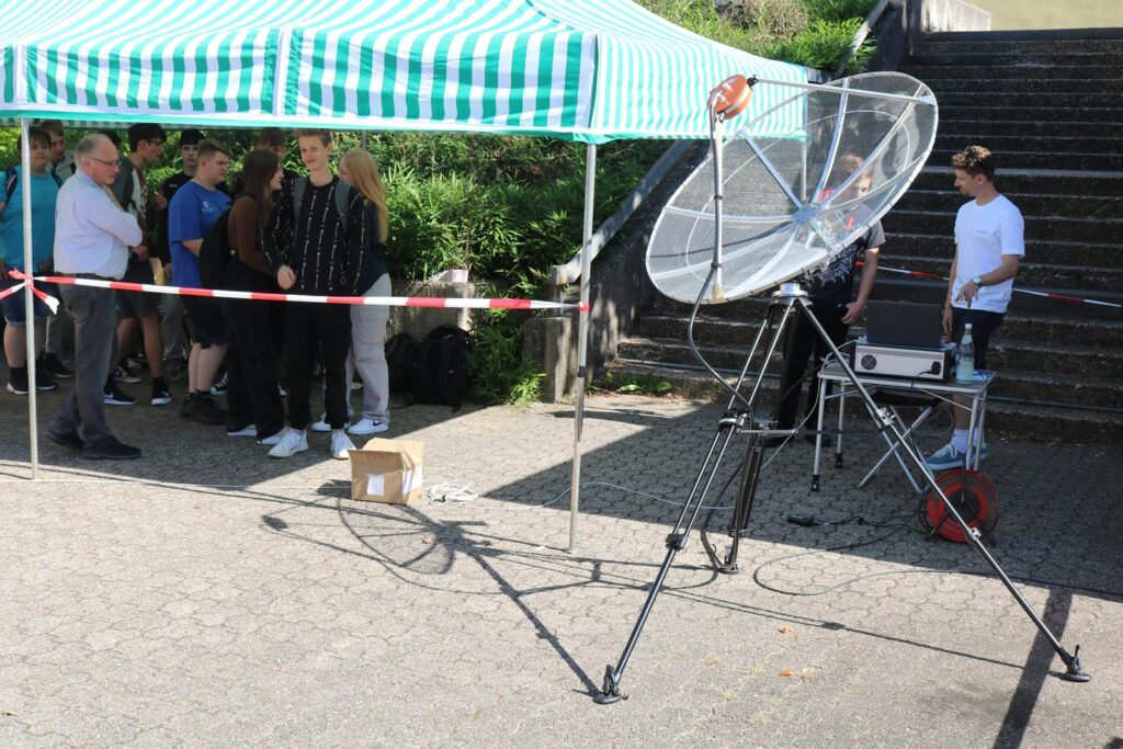 Menschen stehen hinter einer Absperrung unter einem Zeltdach, daneben ein großes Parabolgerät.