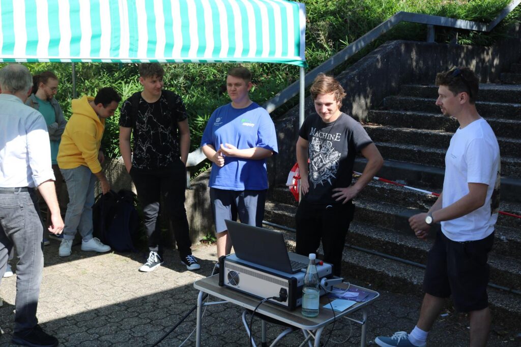 Gruppe von Jugendlichen steht um einen Tisch mit Laptop, im Freien unter einem Pavillon.