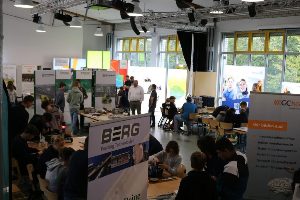 Schulveranstaltung mit Informationsständen und Schülern an Tischen.
