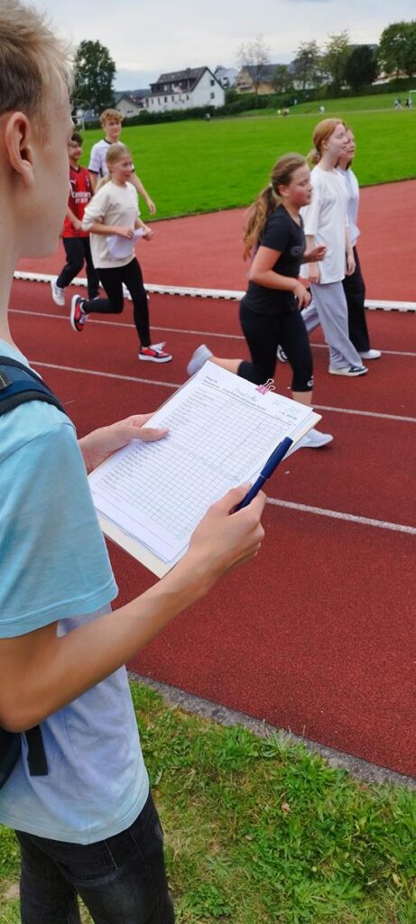 Schüler laufen auf der Laufbahn, ein Mitschüler hält eine Liste in der Hand.