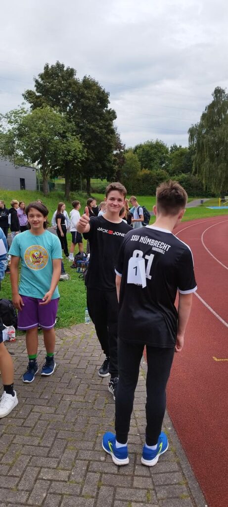 Schüler stehen auf einem Sportplatz, einige lachen und unterhalten sich.