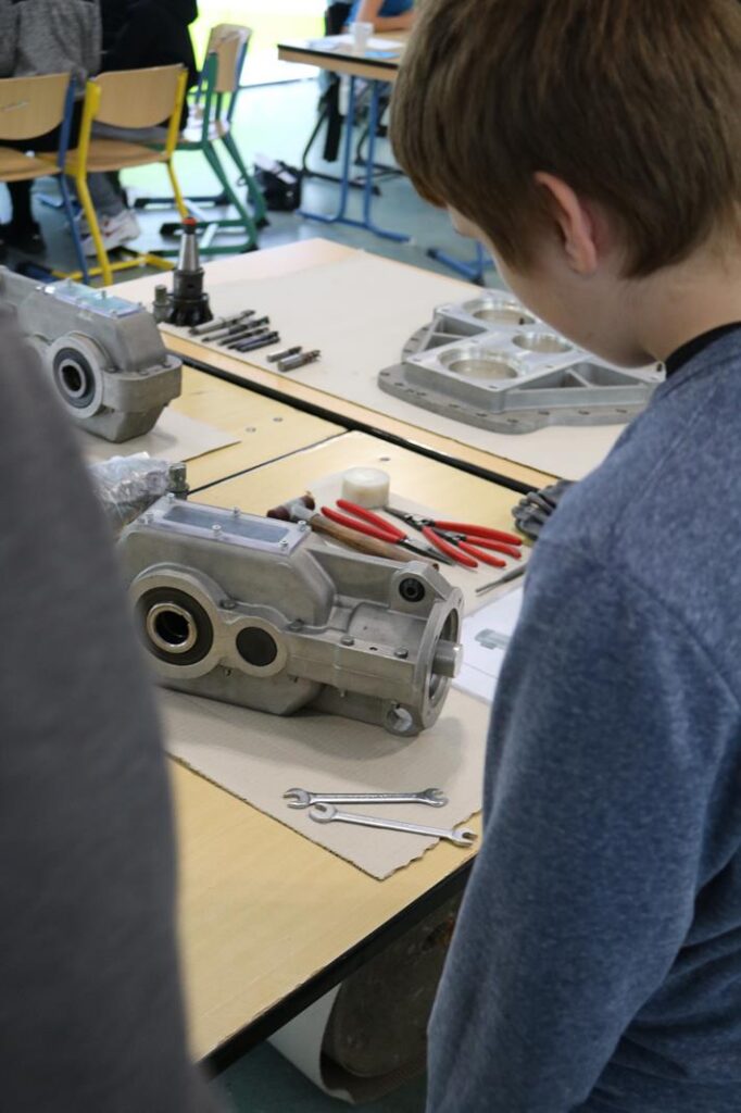 Schüler betrachtet Maschinenbauteile auf einem Tisch in einem Klassenraum.