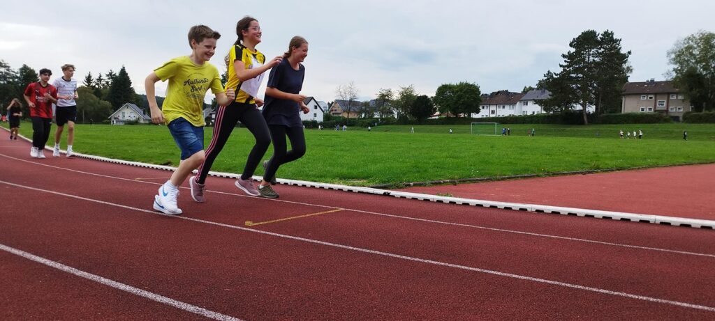 Schüler laufen auf einer Laufbahn, mit Rasen und Bäumen im Hintergrund.