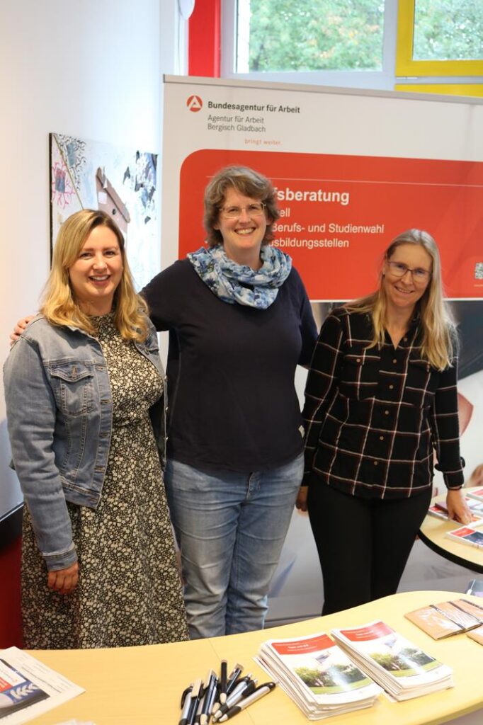 Drei Frauen stehen an einem Informationsstand, umgeben von Broschüren und Stiften.