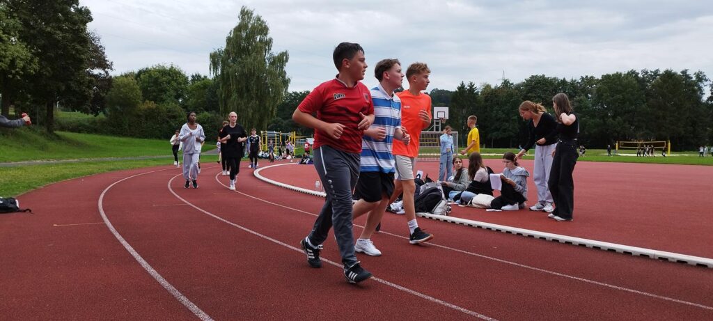 Schüler laufen auf einem Sportplatz, Zuschauer sitzen am Rande.