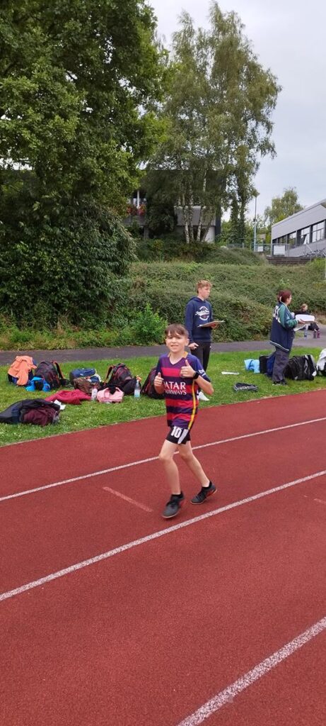 Ein Junge läuft auf einer Leichtathletik-Bahn, im Hintergrund sind Zuschauer und Sportbekleidung.