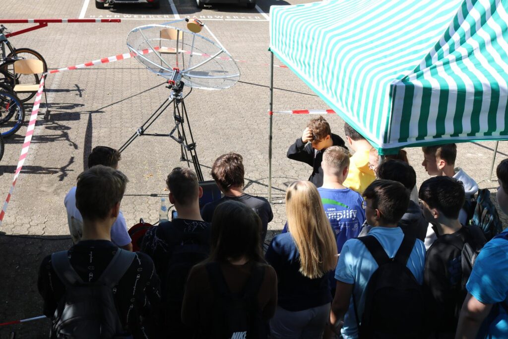 Schüler stehen vor einem Zelt mit einem technischen Gerät und beobachten eine Demonstration.