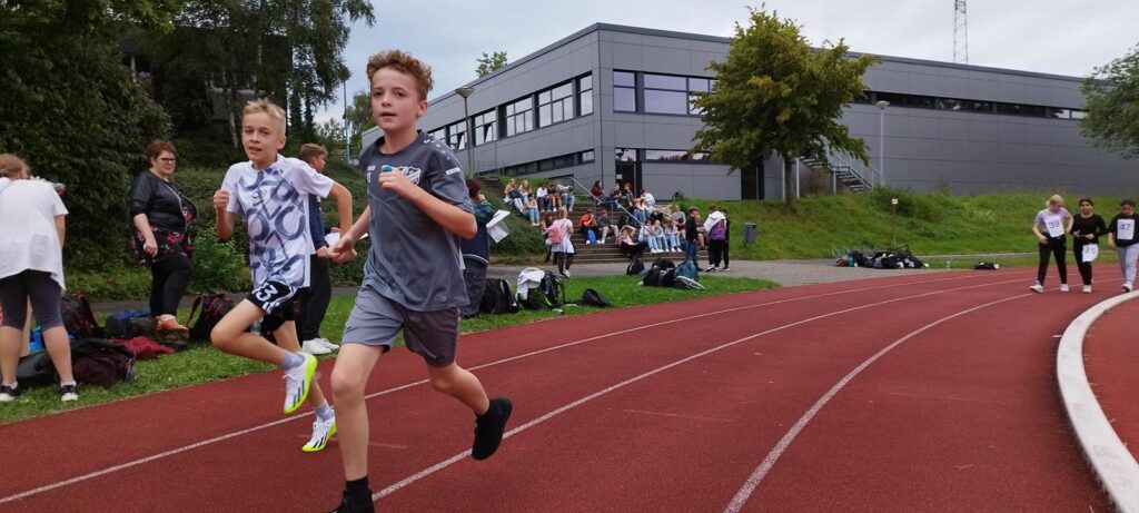 Zwei Schüler laufen auf einer Sportbahn, im Hintergrund steht ein Schulgebäude.