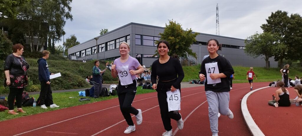 Drei Schülerinnen laufen auf einer Sportbahn, im Hintergrund ein Schulgebäude.