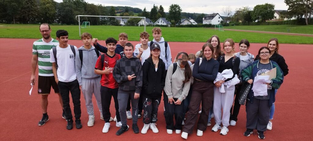 Gruppe von Schülern auf einem Sportplatz, Lehrer steht links.