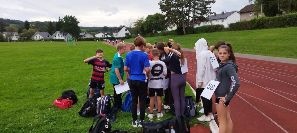 Gruppe von Schülern steht an einer Sportanlage, mit Sporttaschen und Wettkampfnummerschildern.