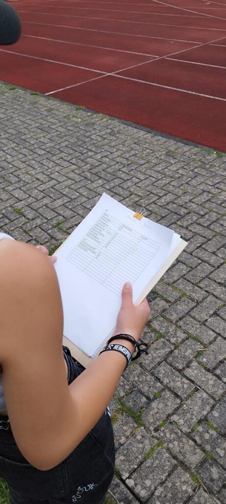 Eine Person hält ein Blatt Papier in der Hand, im Hintergrund ein Sportplatz.