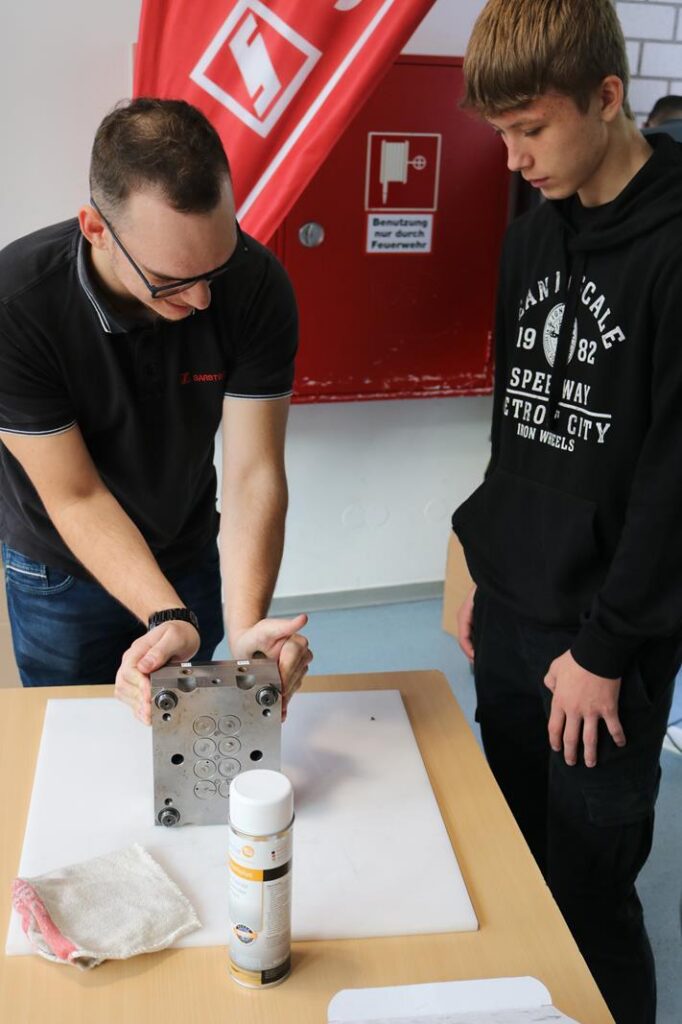 Lehrer zeigt Schüler eine Metallplatte und erklärt praktische Technik.