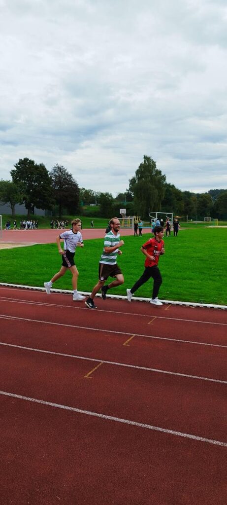 Drei Schüler laufen auf einer Sportbahn unter bewölktem Himmel.