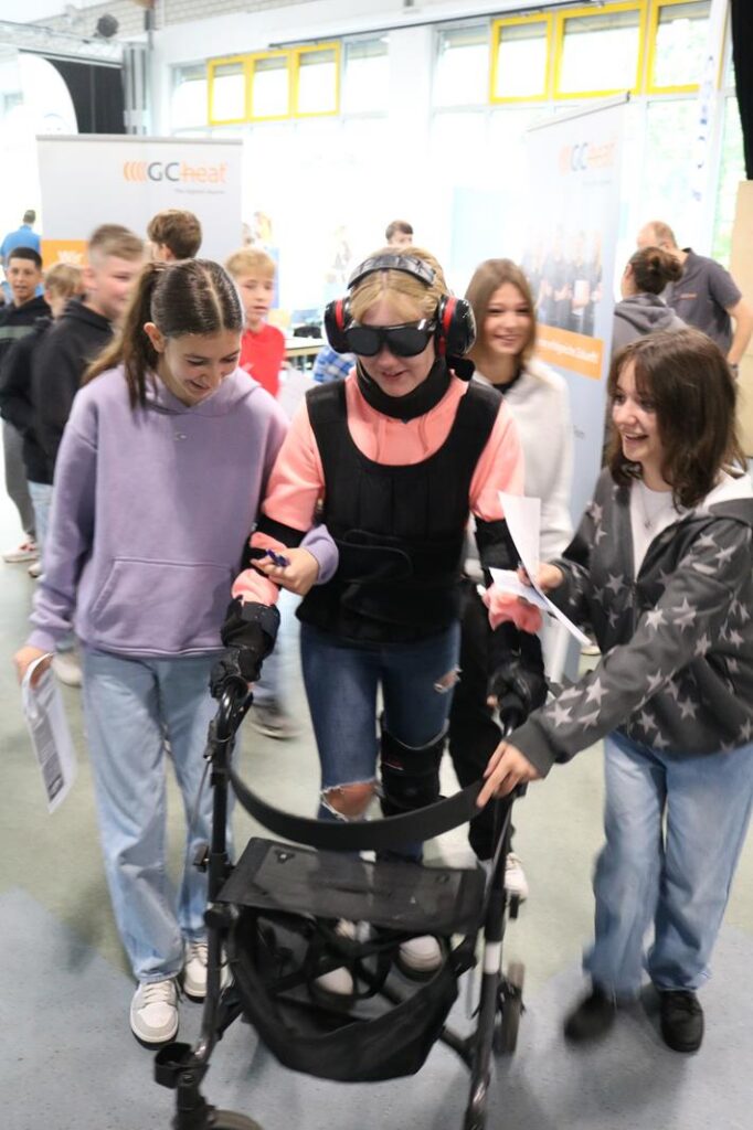 Schülerinnen helfen einer Person mit einem Exoskelett und Rollator.