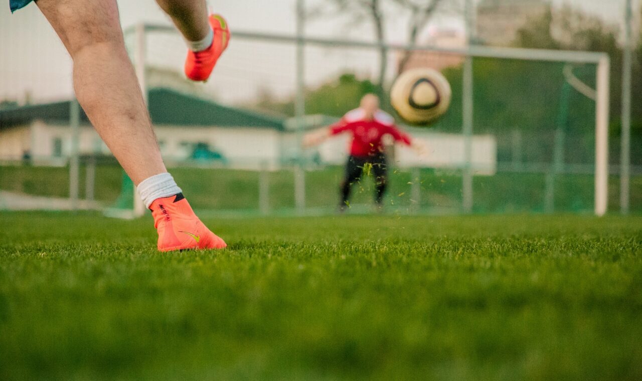 Fußballspieler schießt Ball auf einem Spielfeld, Torwart im Hintergrund.