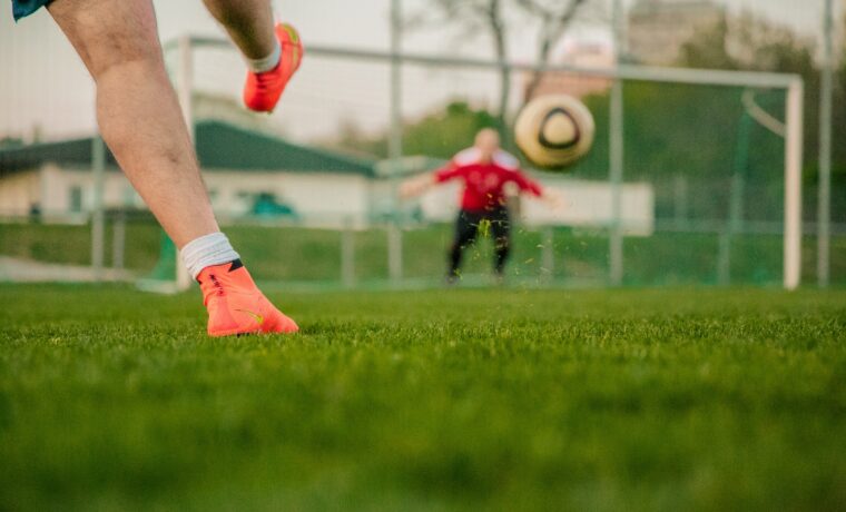 Fußballspieler schießt Ball auf einem Spielfeld, Torwart im Hintergrund.