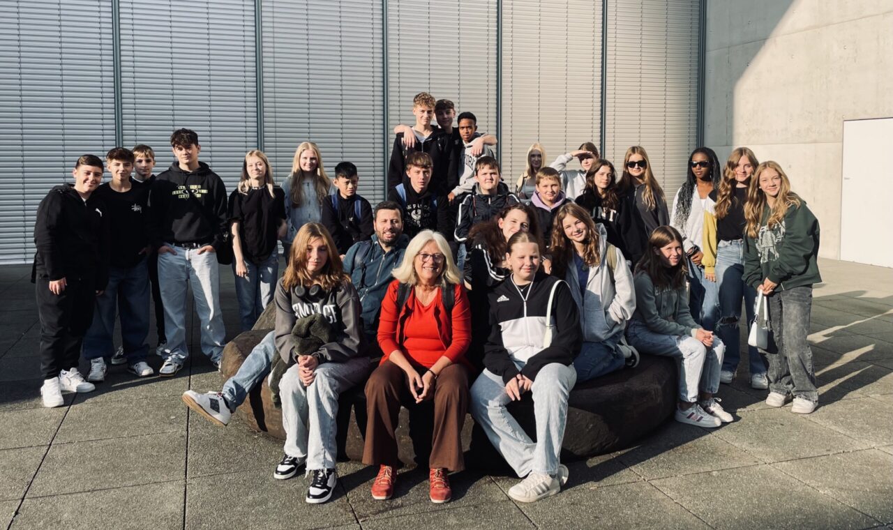 Gruppe von Schülern und Lehrern posiert gemeinsam auf einer Sitzgelegenheit im Freien.