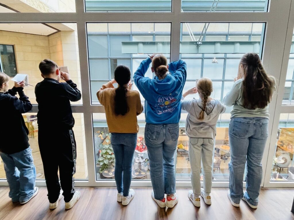 Sechs Kinder stehen am Fenster und schauen nach draußen. Einige machen Fotos.