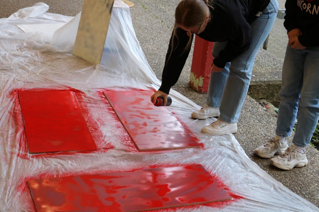 Zwei Personen sprühen rot auf große Platten, die auf einer Plane liegen.