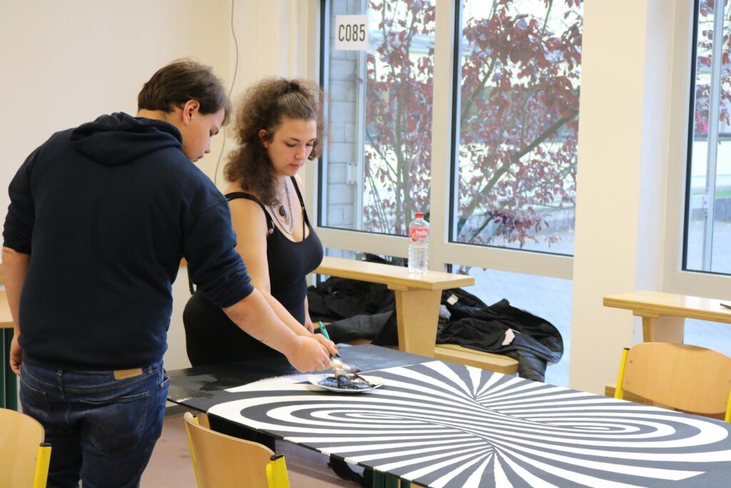 Zwei Personen arbeiten an einem gedrehten Schwarz-Weiß-Muster auf einem Tisch.