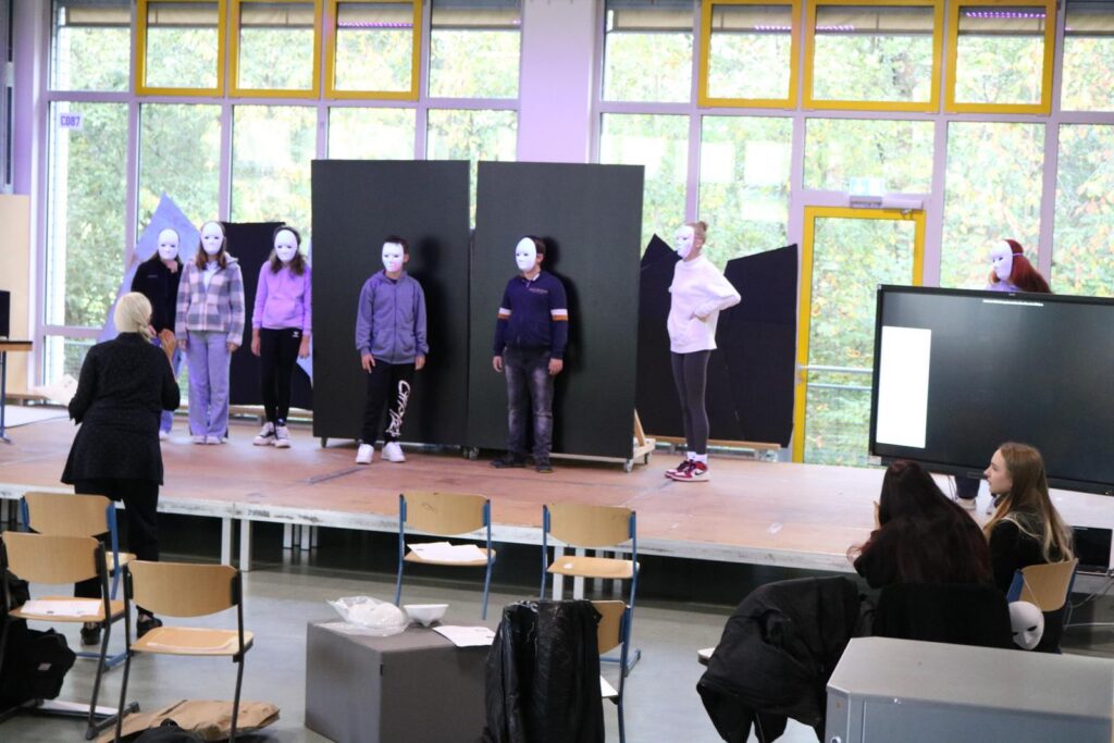 Schüler mit Masken stehen auf einer Bühne in einem Klassenraum.