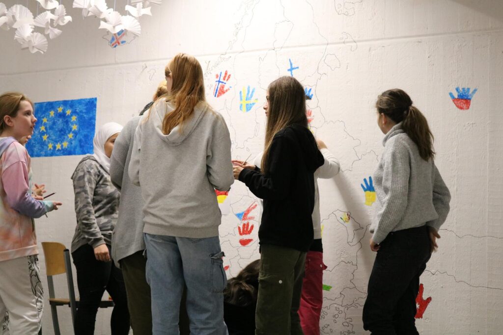 Schülerinnen malen gemeinsam an einer Wand mit bunten Handabdrücken.