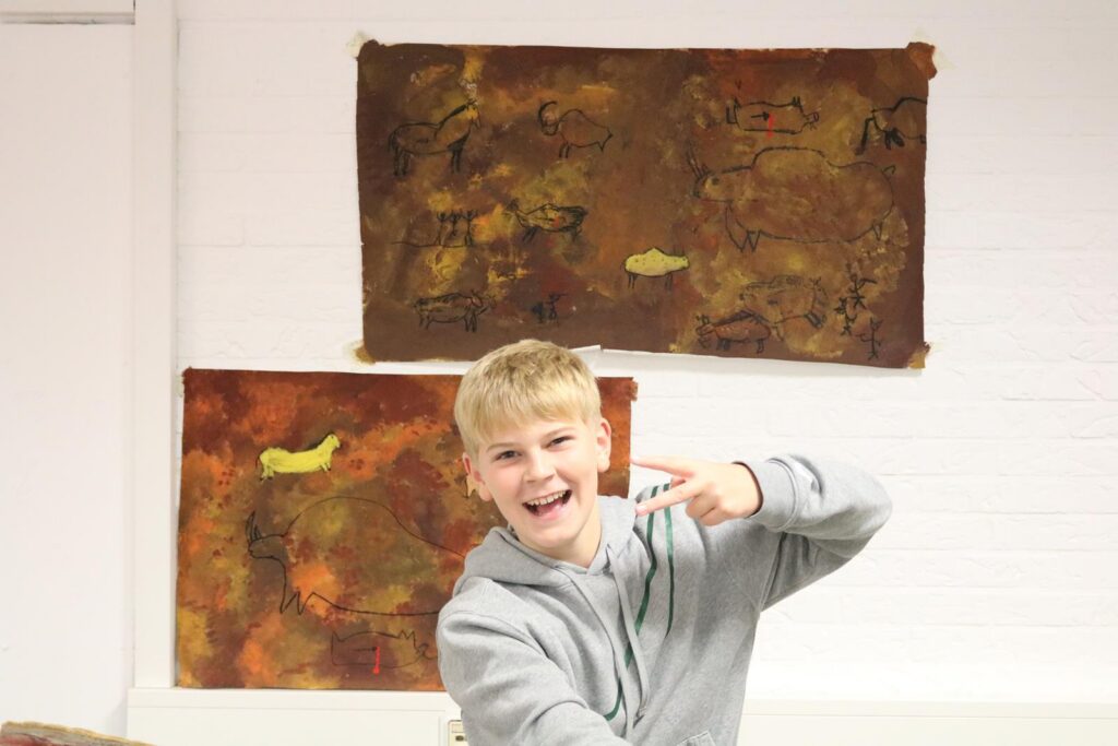 Junge mit kurzen Haaren lächelt und zeigt ein Friedenszeichen vor Kunstwerken an der Wand.