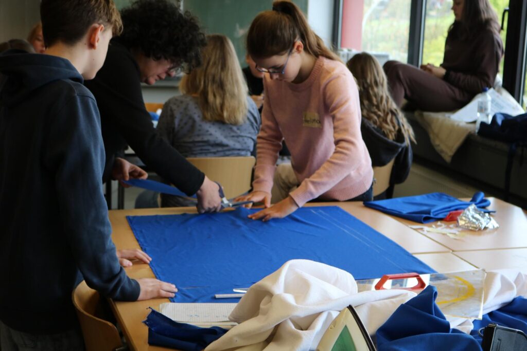 Schüler:innen schneiden blaue Stoffe an einem Tisch in einem Klassenraum zu.