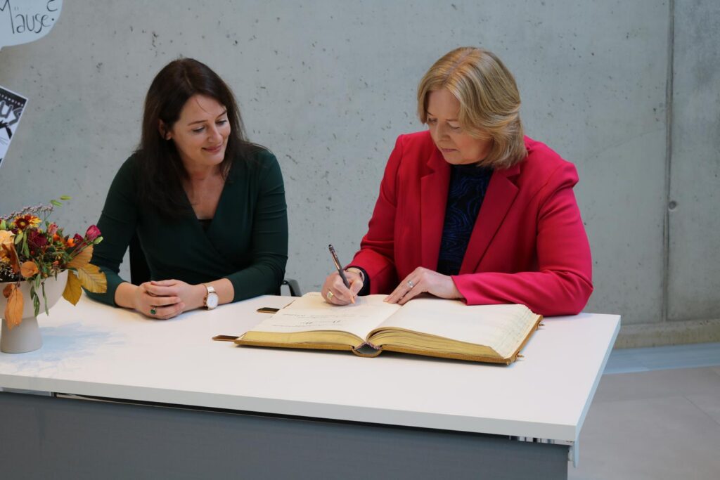 Zwei Frauen sitzen an einem Tisch, eine schreibt in ein großes Buch.