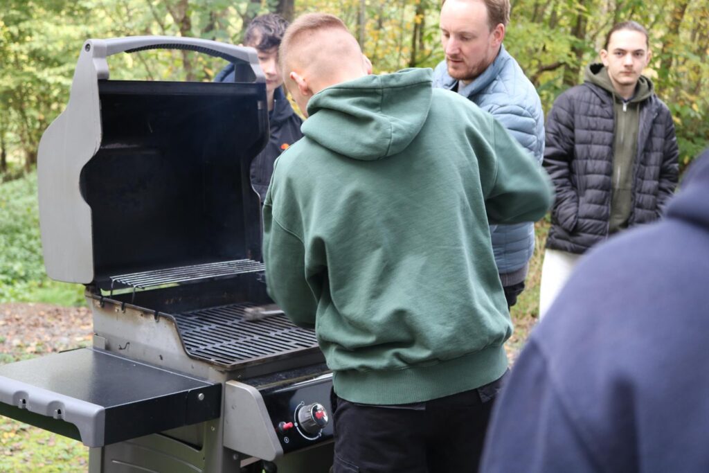 Junge grillt an einem offenen Grill, Zuschauer beobachten im Hintergrund.