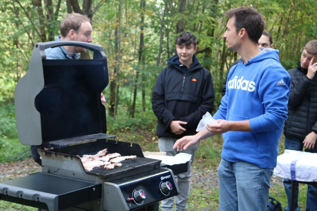 Grillende Personen im Wald, einer wendet Fleisch auf dem Grill.