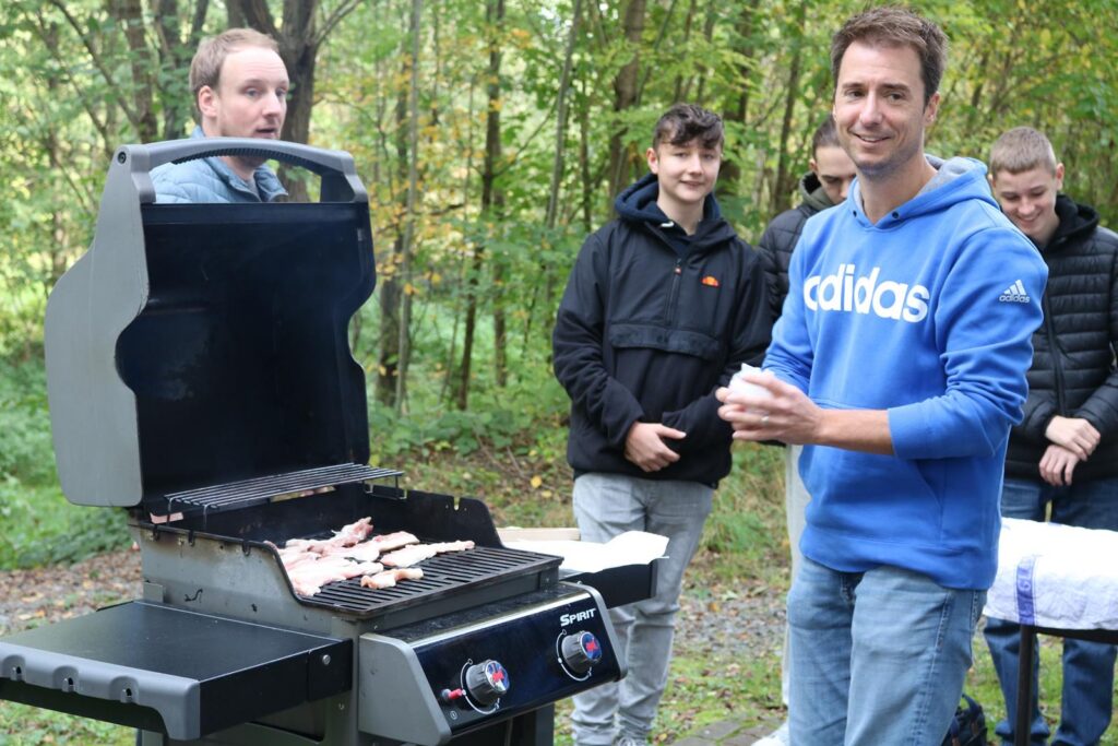 Grillende Personen im Wald mit Speisen auf dem Grill.