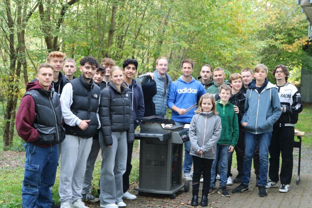 Gruppe von Jugendlichen und Erwachsenen im Wald, steht um einen Grill.