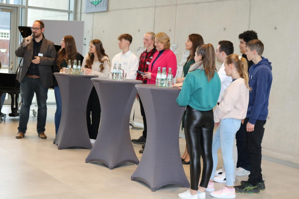 Menschen stehen an Stehtischen und hören einem Redner zu.