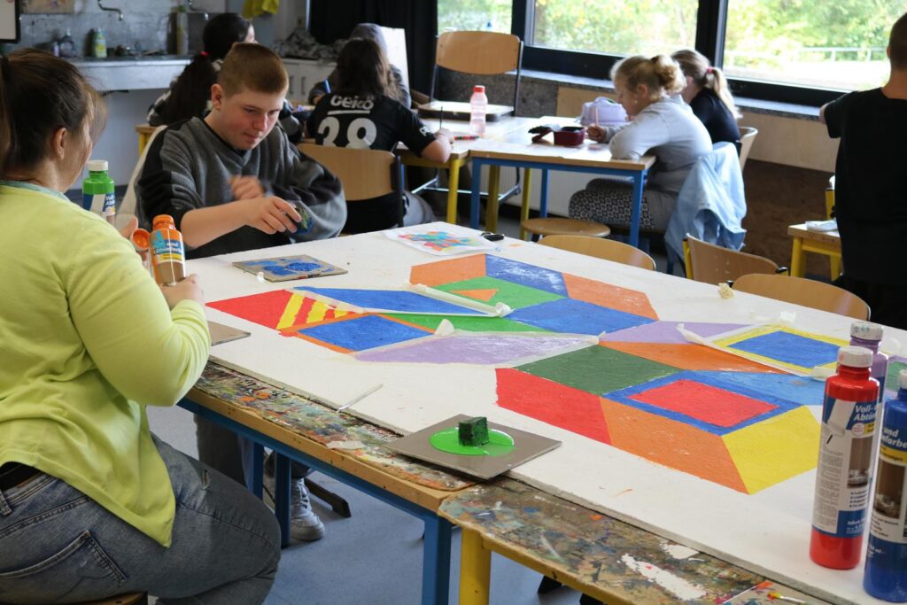 Schüler gestalten ein großes, buntes Kunstprojekt auf einem Tisch in einem Klassenraum.