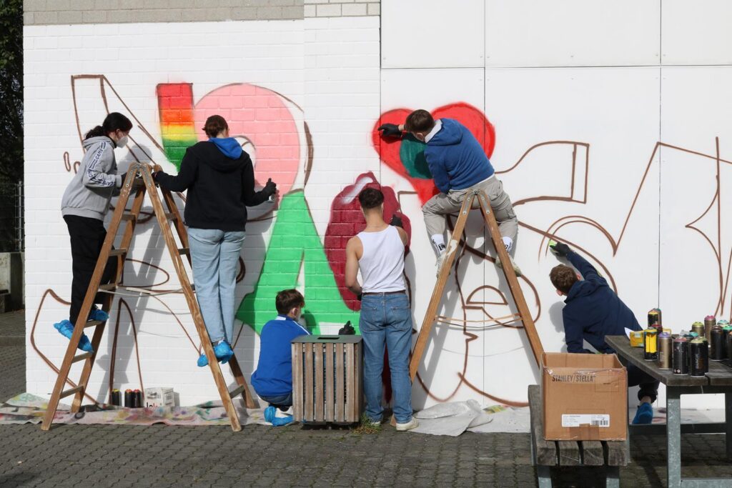 Schüler malen ein Graffiti an einer Wand, stehen auf Leitern und verwenden Farben.