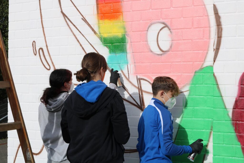 Schüler sprayen ein buntes Wandgemälde auf einer weißen Fläche.