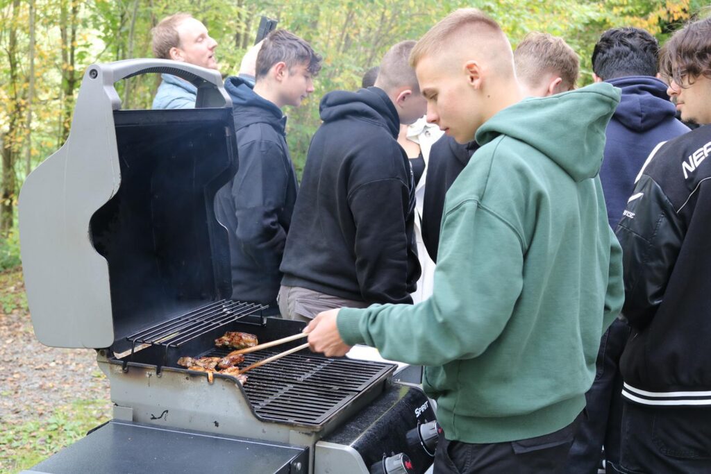 Schüler grillen im Freien, einer hält eine Zange in der Hand.