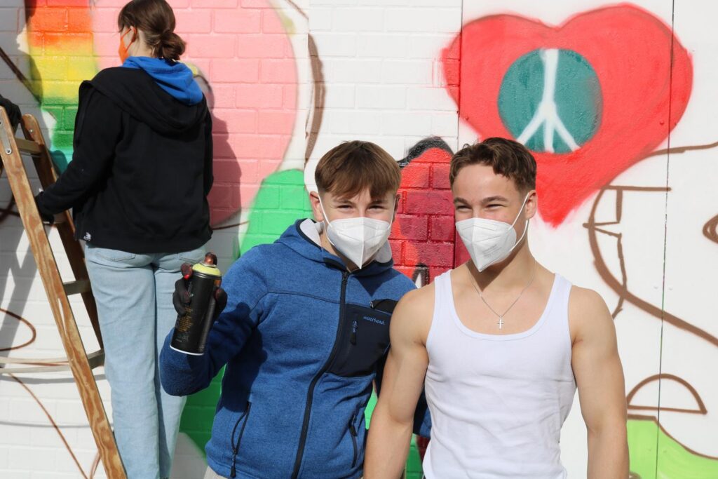 Zwei Jugendliche posieren vor einer Wand mit Graffiti, im Hintergrund malt jemand.