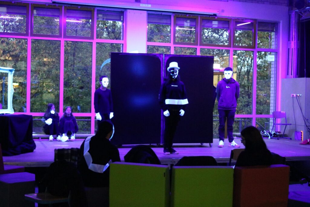 Schüler in Theaterkostümen führen vor einer Kulisse auf. Bunte Beleuchtung im Hintergrund.