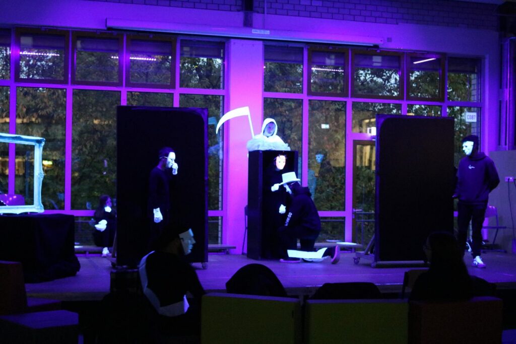 Theateraufführung mit schwarzen Kostümen und Masken, beleuchtet in lila Licht.